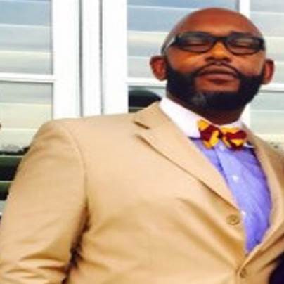 Man in tan suit and bow tie standing confidently.