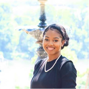 Smiling woman in black dress with pearl necklace outdoors near a fountain.